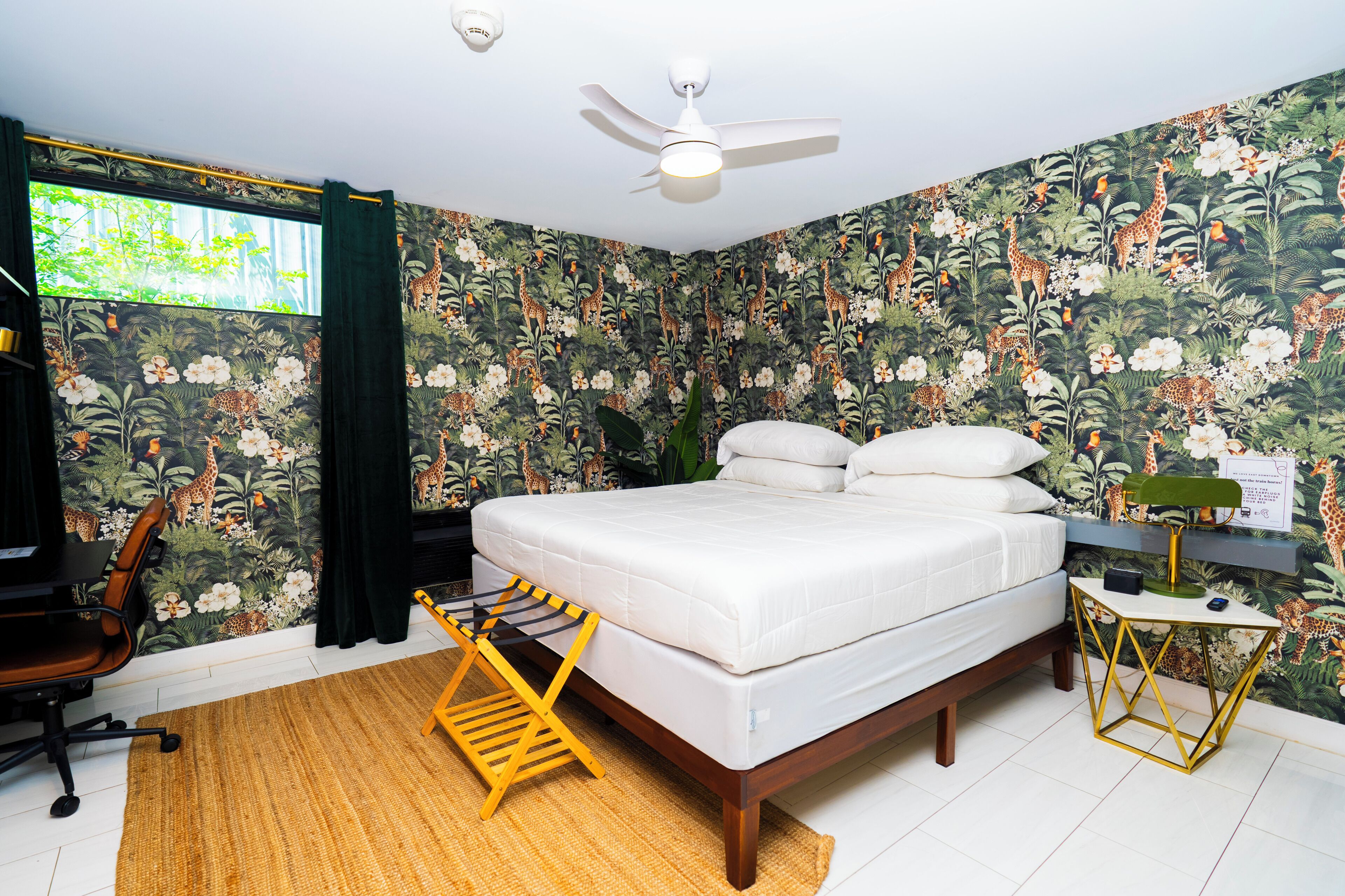A bedroom with a bed, a desk, a chair, a standing fan, and a window with curtains.