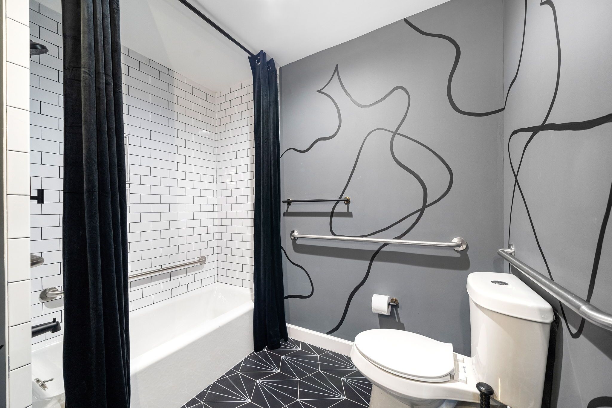 Une salle de bain moderne avec une baignoire blanche, des carreaux de sol noirs à motifs géométriques, des toilettes blanches et un rideau foncé.