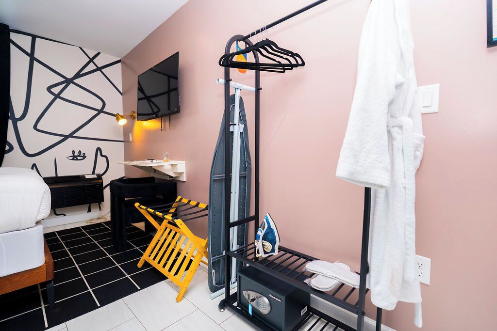 A hotel room with a bed, a black and white checkered floor, a black iron, a yellow laundry basket, and a white towel hanging on a rack.