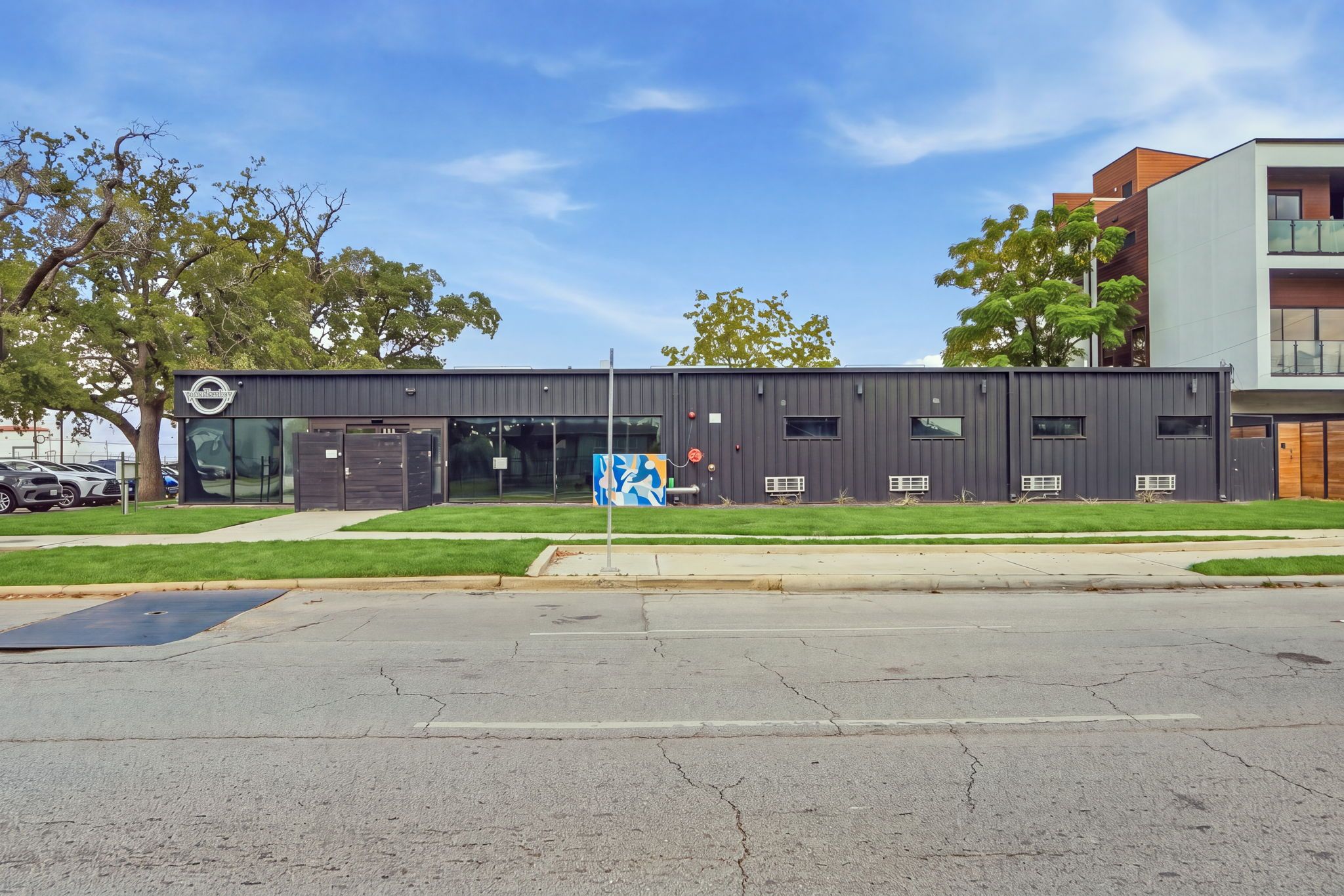 A modern building with a flat roof, large windows, and a parking lot in front.