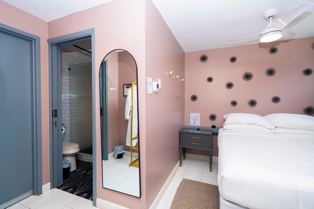 Une chambre à coucher comprenant un lit, une table de chevet, un miroir, un ventilateur de plafond, ainsi qu’une salle de bain avec des toilettes et un lavabo.