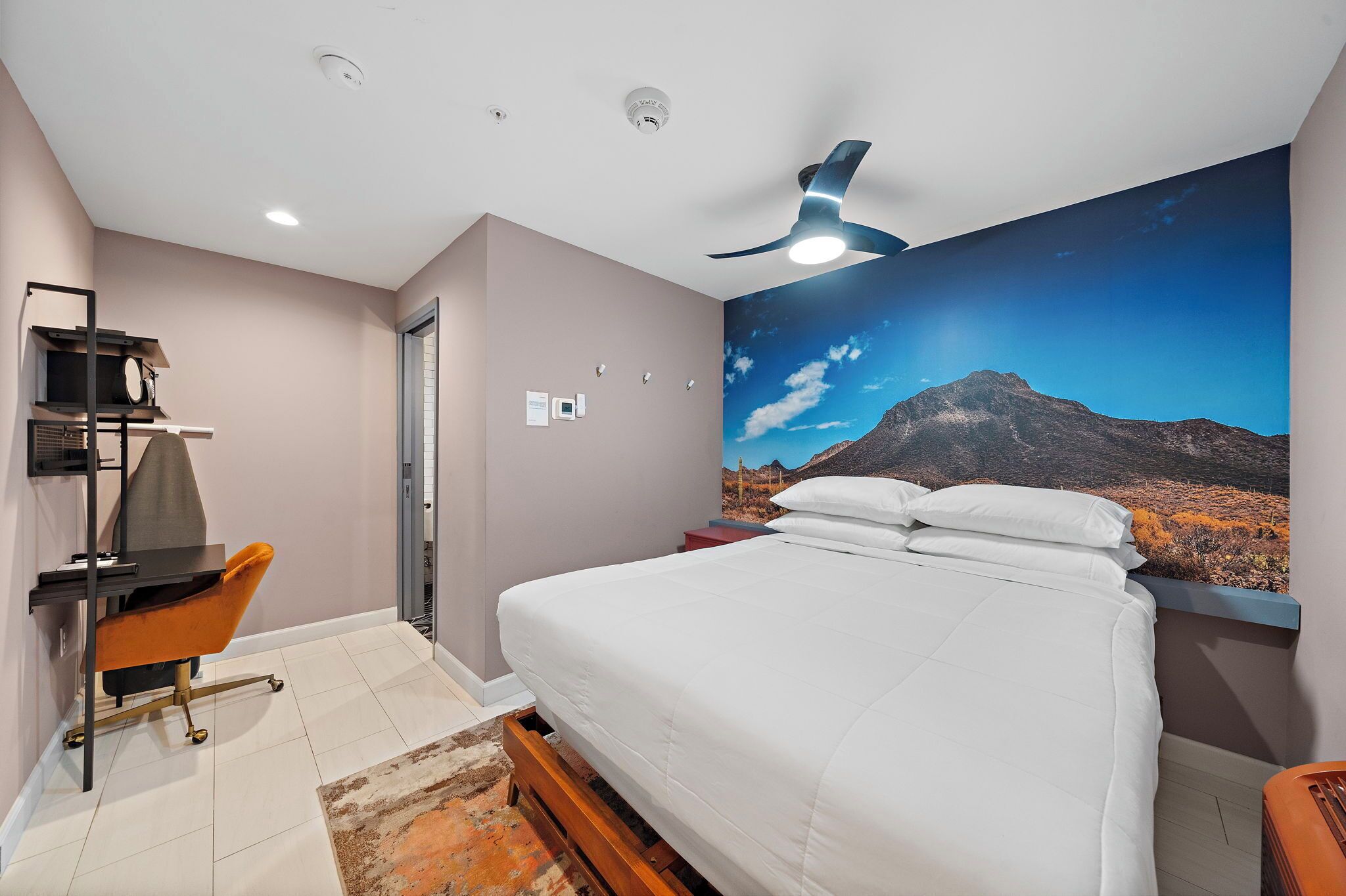 A bedroom with a bed, a desk with a chair, a ceiling fan, and a wall-mounted TV.