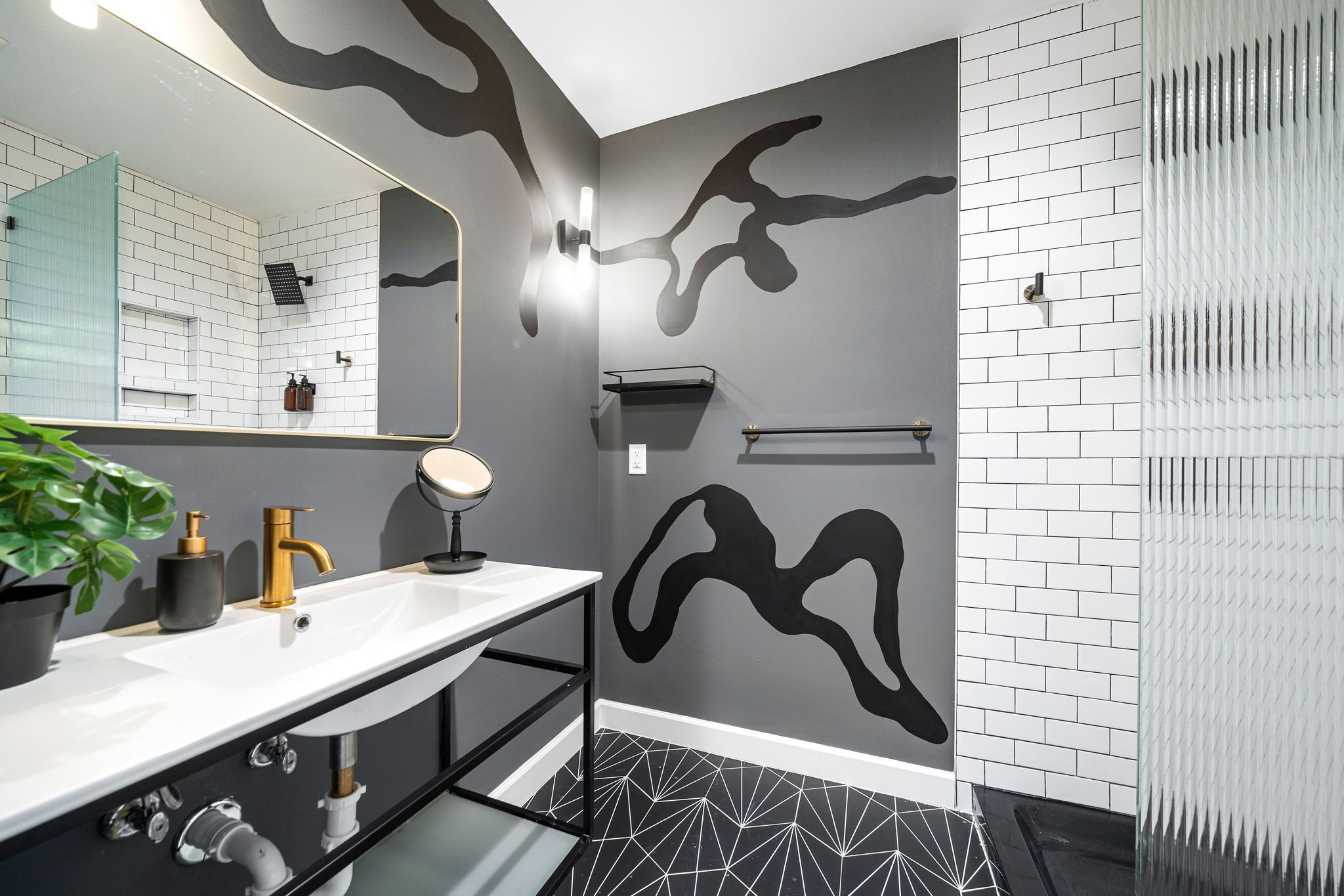 A modern bathroom with a white sink, gold faucet, and a large mirror. There is a black abstract wall design and a patterned floor.