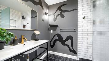 A modern bathroom with a white sink, gold faucet, and a large mirror. There is a black abstract wall design and a patterned floor.