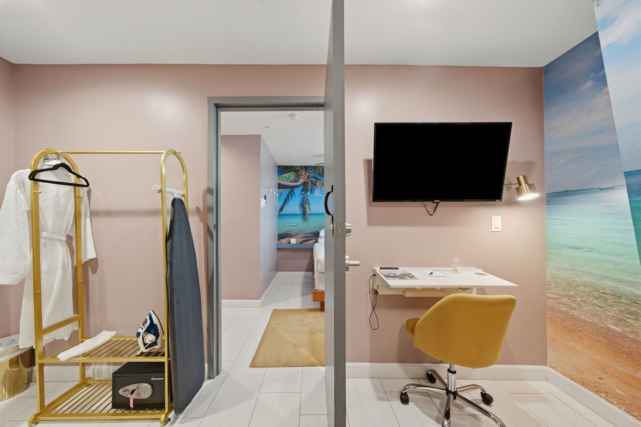 Habitación de hotel con cama, escritorio con silla, televisor en la pared y baño con tendedero y tabla de planchar.