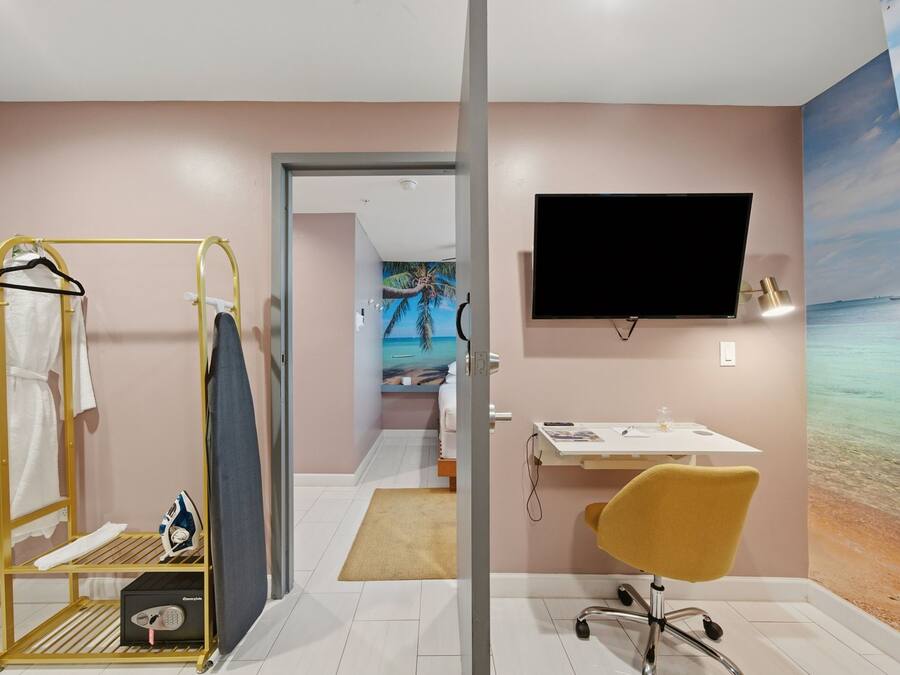Habitación de hotel con cama, escritorio con silla, televisor en la pared y baño con tendedero y tabla de planchar.