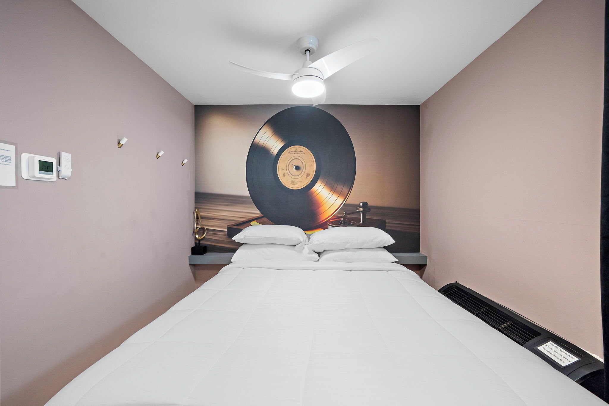 A hotel room with a bed, a ceiling fan, a wall-mounted clock, and a large vinyl record on the wall.