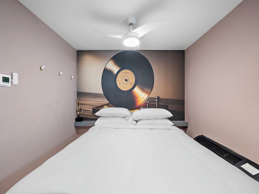 A hotel room with a bed, a ceiling fan, a wall-mounted clock, and a large vinyl record on the wall.