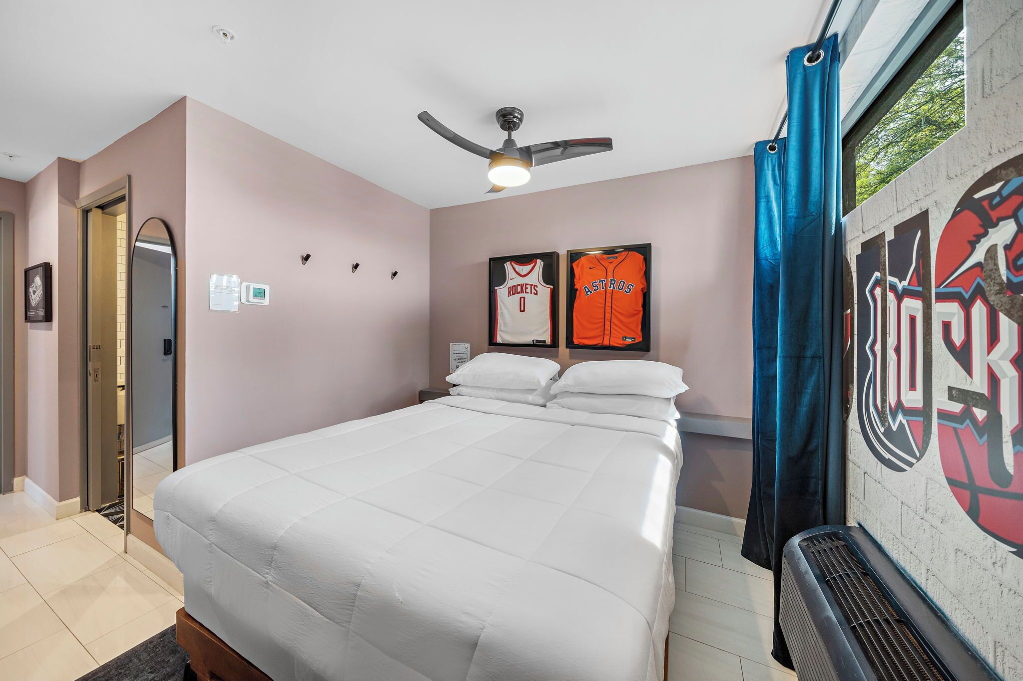A hotel room with a bed, a ceiling fan, a window with curtains, and sports memorabilia on the wall.