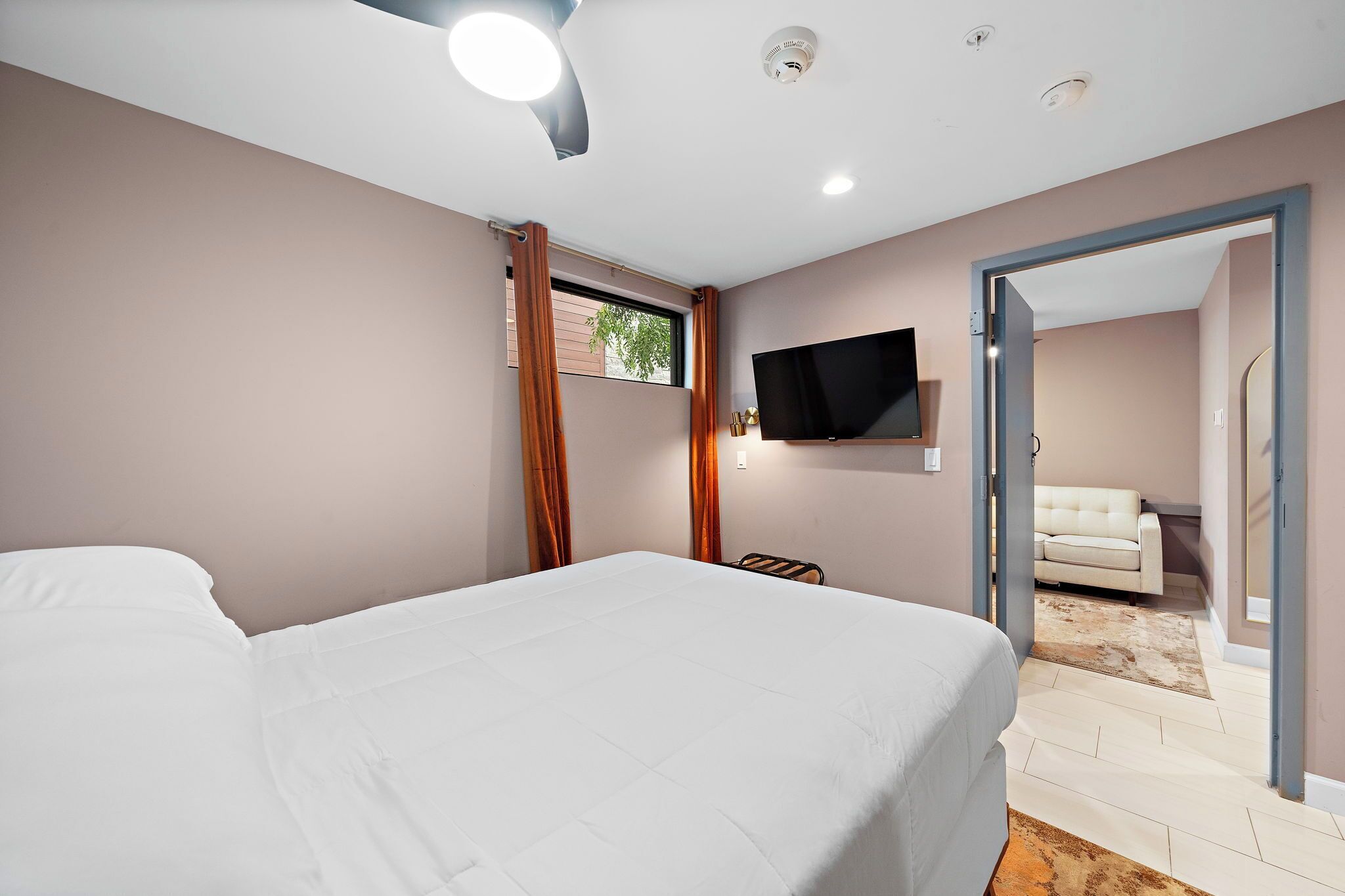Un dormitorio con cama, televisor montado en la pared y vista a un salón a través de una puerta abierta.