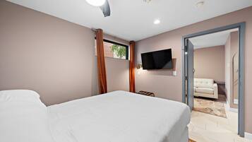 Un dormitorio con cama, televisor montado en la pared y vista a un salón a través de una puerta abierta.