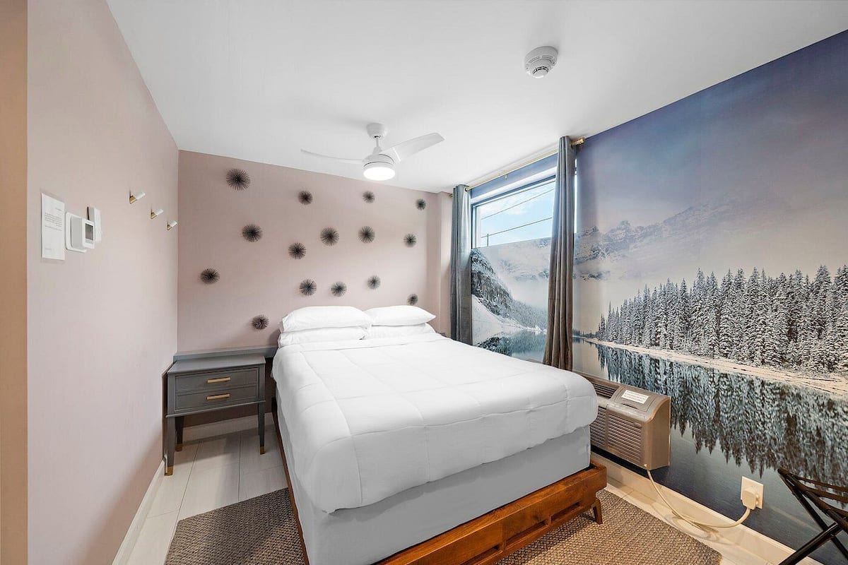 Een slaapkamer met een bed, een nachtkastje, een wandheater, een plafondventilator en een groot raam met uitzicht op de bergen.