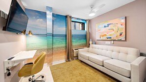 Quarto com escrivaninha, sofá, televisão, ventoinha no teto e um mural com tema de praia.