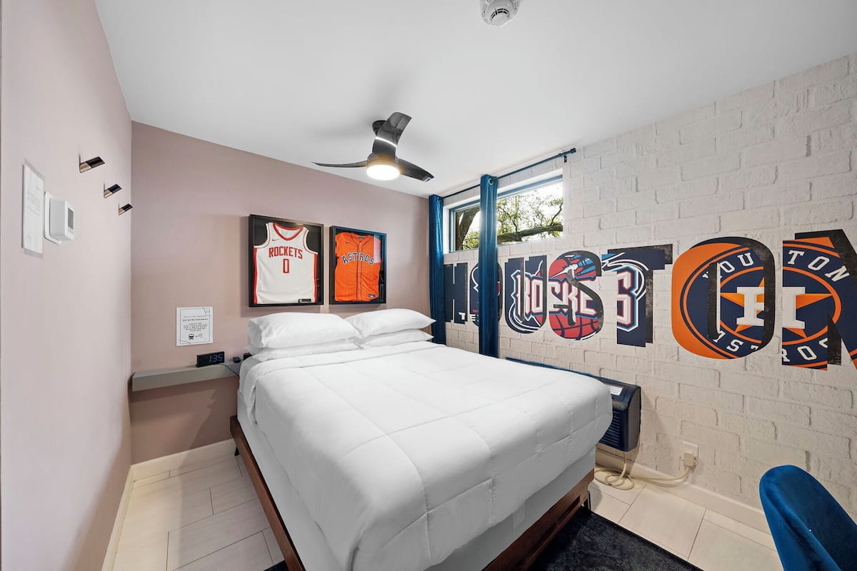 Een slaapkamer met een bed, een plafondventilator, een wandklok en sporttenue aan de muur.