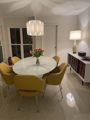 A dining area with a round table, yellow chairs, a vase with flowers, a pendant light, and a cabinet with a lamp.