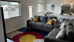 Ein modernes Wohnzimmer mit einem grauen Sofa, einem roten und gelben Teppich und einem Küchenbereich mit Edelstahlgeräten.