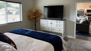 Ein Schlafzimmer mit Bett, Kommode, Fernseher, Pflanze und Blick ins Freie.
