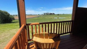 Una terraza de madera con una silla de madera, vista a un campo cubierto de hierba y un edificio distante.