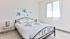 A neatly made bed with white linens and pillows, a bedside table with a lamp, and a wall with floral decals. - Le Gois - Jardin -  Marais salants (Beauvoir-sur-Mer)