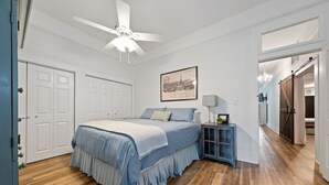 A bedroom with a bed, a ceiling fan, a nightstand, a framed picture, and a door leading to another room. - RoosterHouse Countryside Pecan Farm 6ac SanMarcos (Maxwell)