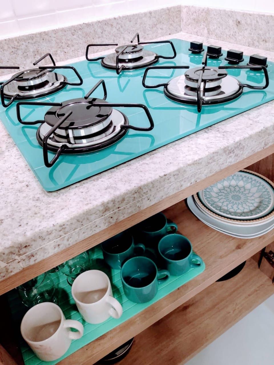 Uma cozinha com fogão a gás turquesa, prateleira de madeira com canecas e bancada de granito.