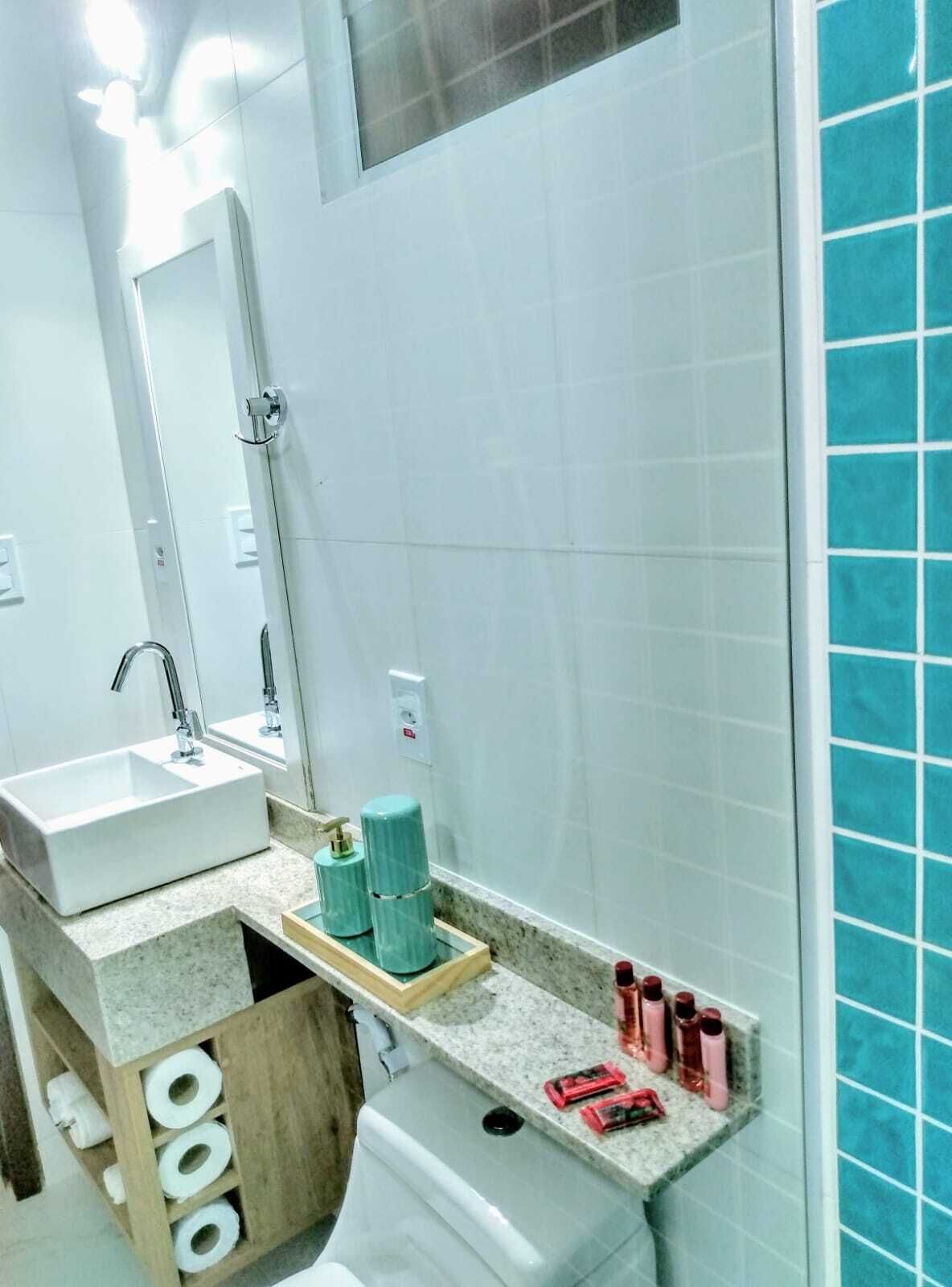 Casa de banho com lavatório, espelho e sanita. Artigos de higiene pessoal estão no balcão e uma área de duche com azulejos azuis.