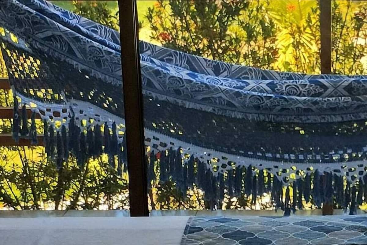 A hammock with a patterned cover, a thatched roof structure, and a view of a lush garden with various plants and a hill in the background.