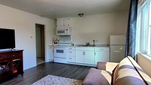Ein modernes Wohnzimmer mit Flachbildfernseher, einer Holz-Unterhaltungsecke und einer Couch. Im Hintergrund ist eine Küche mit weißen Schränken, einer Mikrowelle und einem Kühlschrank zu sehen.