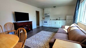 Ein Wohnzimmer mit einer Couch, Sesseln, einem Fernseher, einem Esstisch und einer Küchenzeile mit Schränken und Geräten.