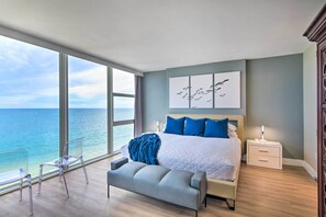 Ein Schlafzimmer mit großem Fenster und Meerblick, einem Bett mit blauen Kissen, einem Nachttisch und einer Bank.