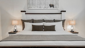 Una cama bien hecha con una colcha blanca y varios almohadones, flanqueada por dos mesitas de noche con lámparas. Un cuadro grande de un bisonte en un bosque cuelga sobre la cama.