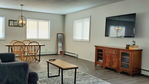 Un salon comprenant un canapé, une table à manger avec des chaises, un téléviseur fixé au mur et un meuble en bois.
