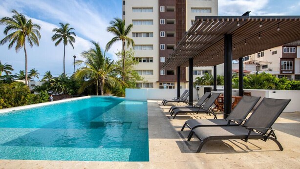 A swimming pool with lounge chairs and palm trees, surrounded by a building with multiple floors and balconies.