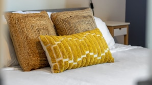 Cama bem arrumada com duas almofadas texturizadas, luminária fixada na parede e uma pequena mesa de cabeceira.
