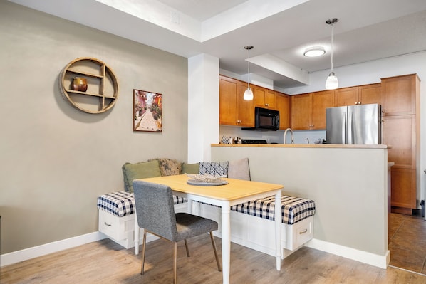 A compact kitchen with wooden cabinets, a stainless steel refrigerator, and a dining area with a table and bench.