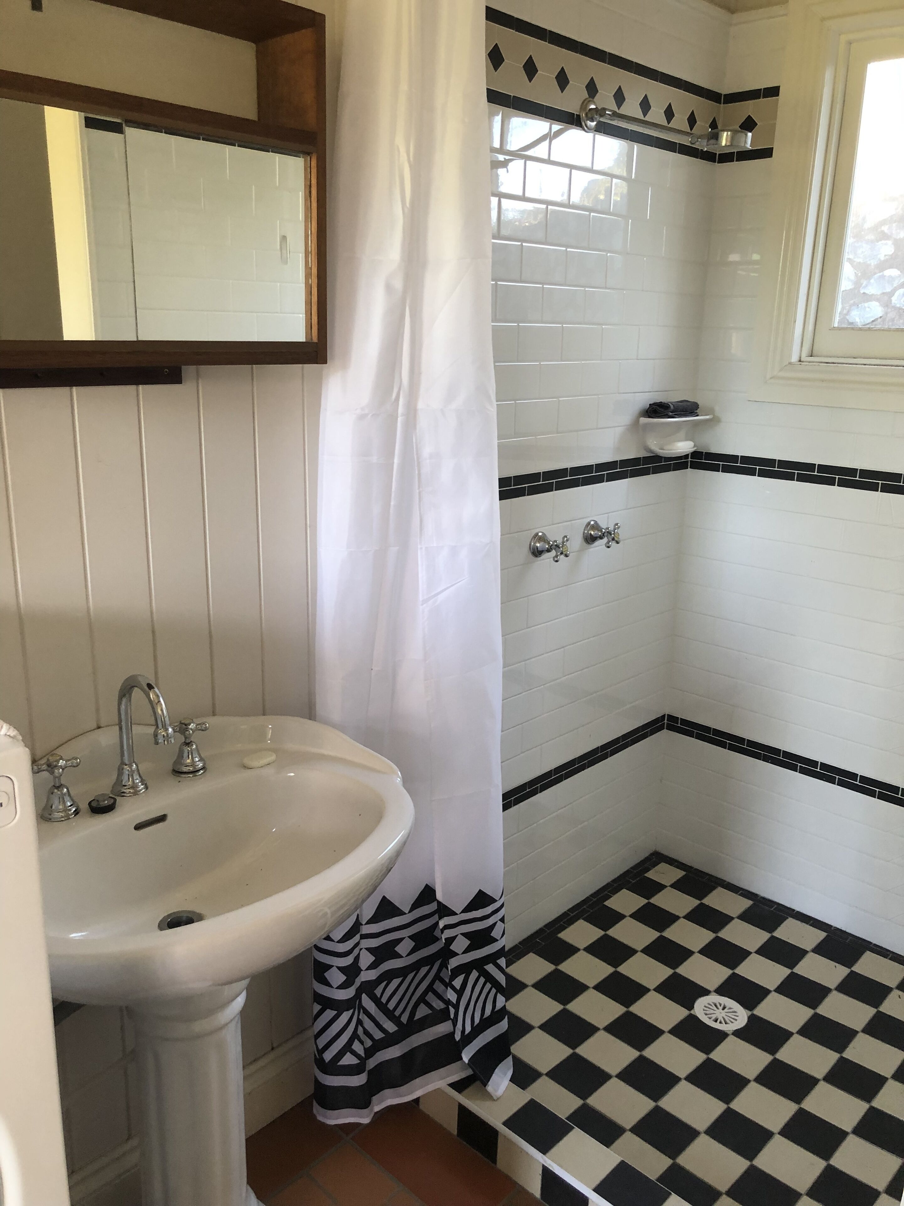Een badkamer met een witte wastafel, een spiegel, een douche met een zwart-wit schaakbordvloer en een wit gordijn.