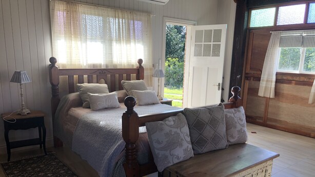 A bedroom with a wooden bed, a wooden bench, a bedside table with a lamp, and a view of the outdoors through a door. - Country Lane Cottage (Mount Samson)