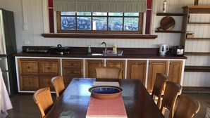 A wooden dining table with chairs, a kitchen with wooden cabinets, a window with red and green patterned glass, and a shelf with kitchen items. - Country Lane Cottage (Mount Samson)