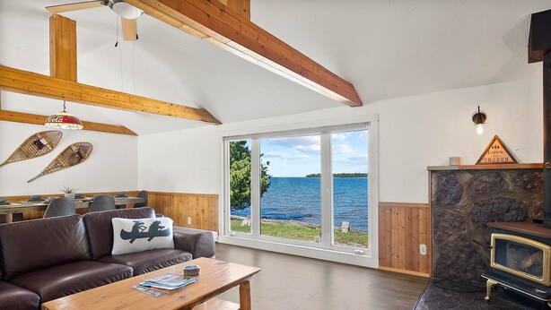Un salon chaleureux avec un plafond en bois, une cheminée en pierre et une grande fenêtre donnant sur un plan d’eau.