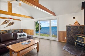 Un salon chaleureux avec un plafond en bois, une cheminée en pierre et une grande fenêtre donnant sur un plan d’eau.