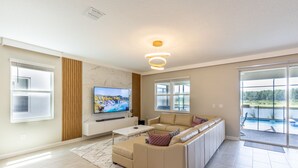 A modern living room with a large L-shaped sofa, a flat-screen TV mounted on the wall, and a sliding glass door leading to a balcony.