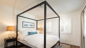 A four-poster bed with a white bedspread and pillows, flanked by two bedside tables with lamps, a framed picture on the wall, and a window with shutters.