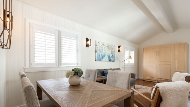 A dining area with a wooden table, chairs, and a vase of flowers. There are windows with blinds, a painting on the wall, and a cabinet in the background.