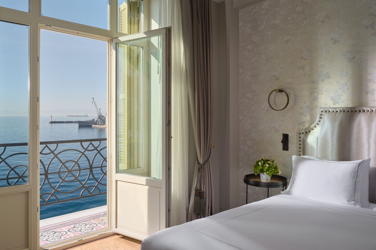 A hotel room with a bed, a small table with a plant, a view of the sea, and a balcony with a railing.