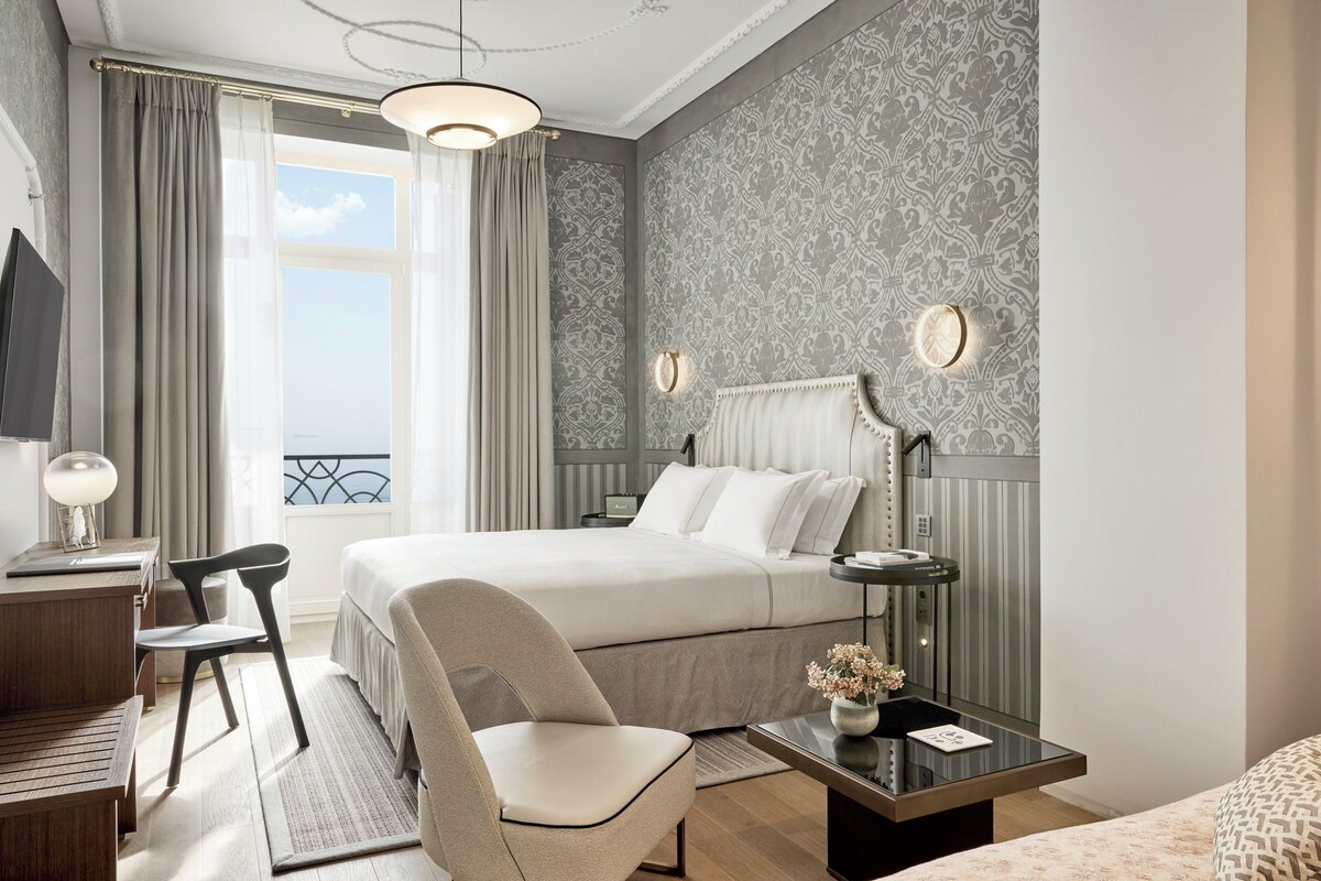 A hotel room with a bed, a desk with a chair, a bedside table, and a view of the outdoors.