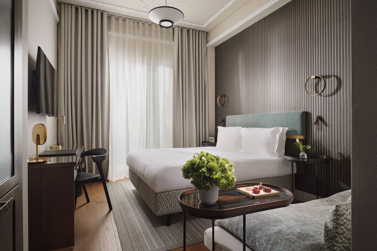 Une chambre d’hôtel comprenant un lit, un bureau, une chaise, une télévision, une table de chevet avec une plante et une fenêtre avec des rideaux.