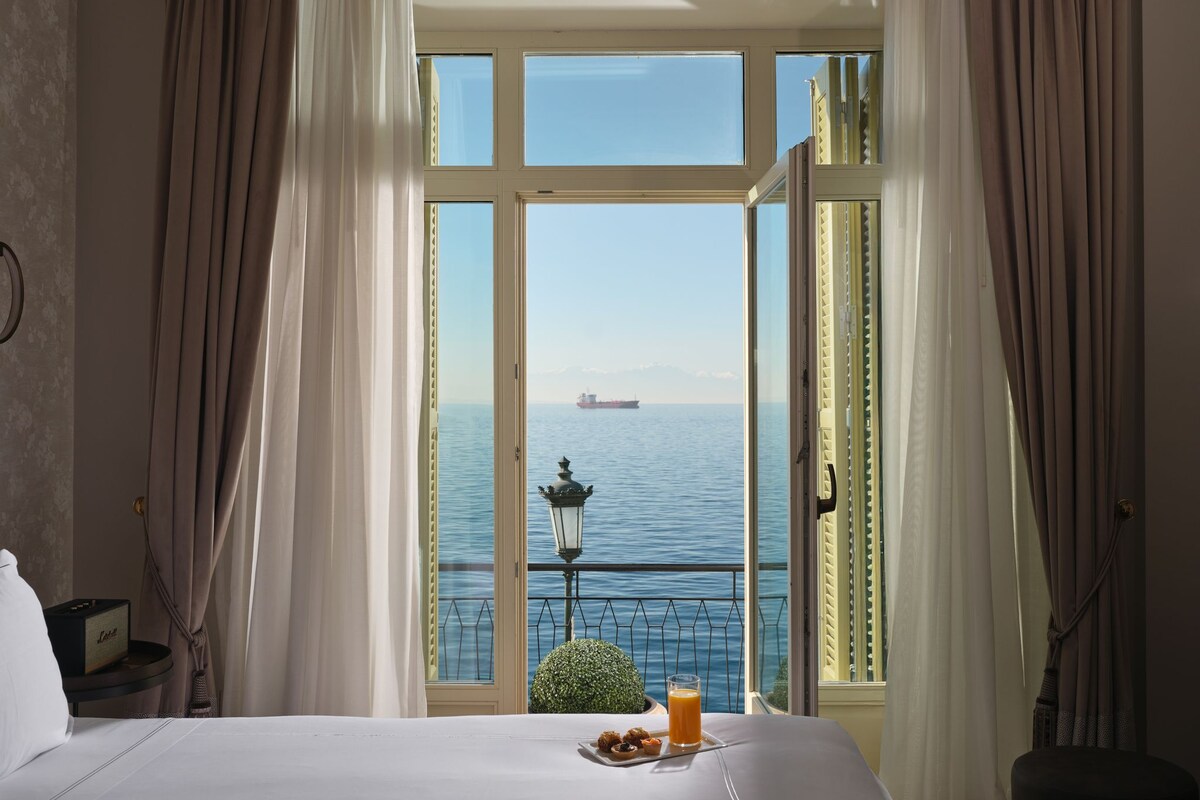 A hotel room with a large window, a bed, a small table with breakfast, and a view of the sea.