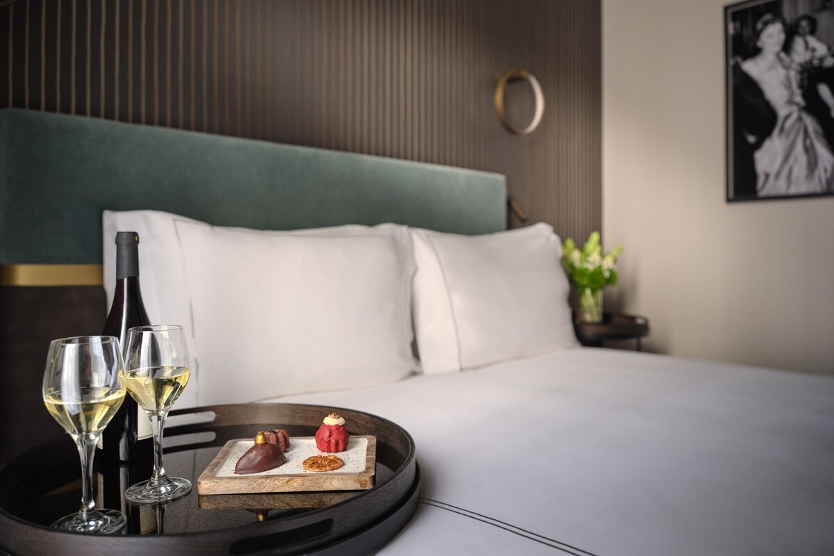 A neatly made bed with white linens, a bedside table with a bottle and glasses, and a tray with desserts.