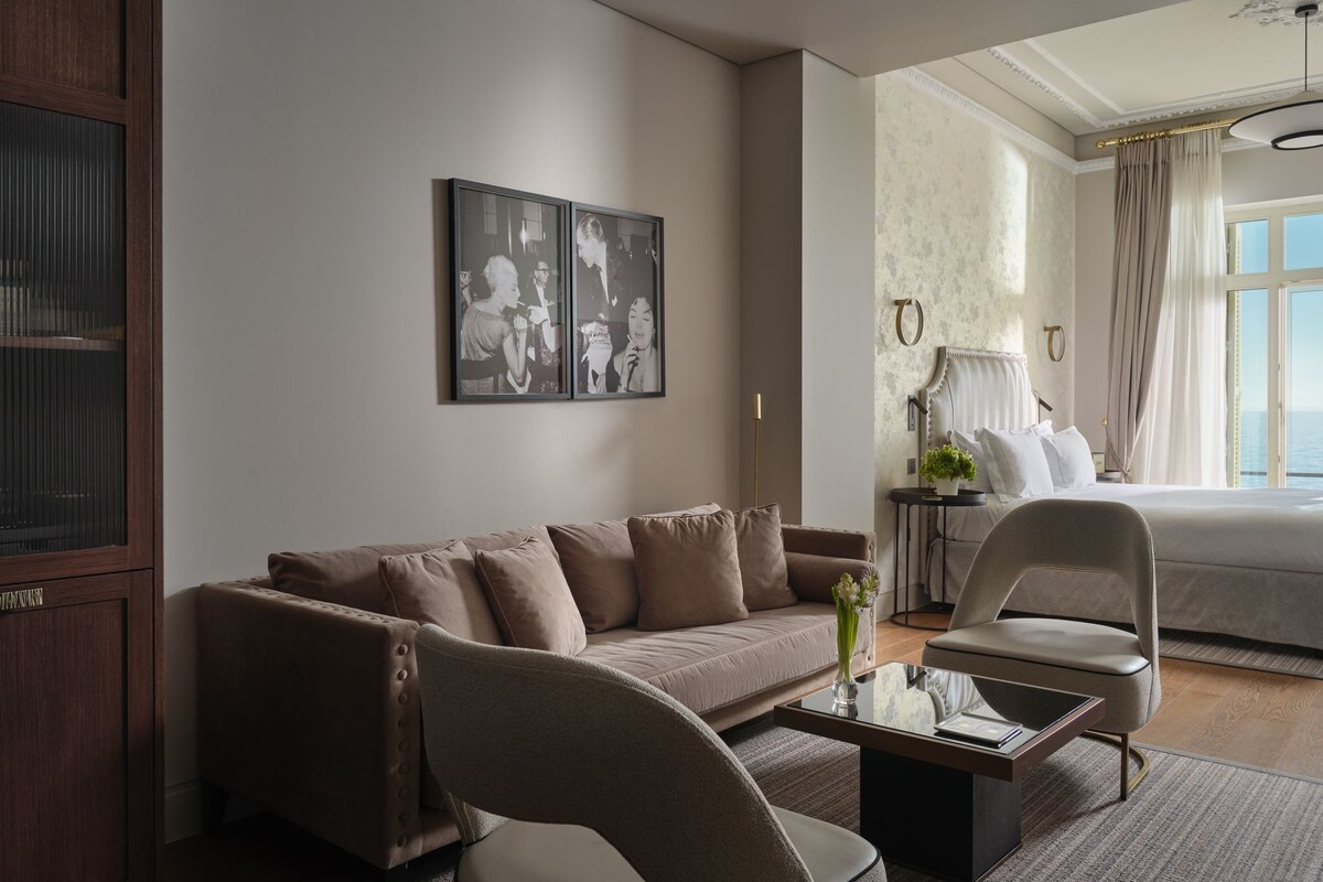 A hotel room with a sofa, armchair, and a bed. There is a framed picture on the wall and a window with curtains.