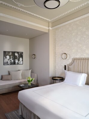 Een moderne hotelkamer met een groot bed, een bank, een kleine tafel met een vaas, en lijstjes met foto’s aan de muur.